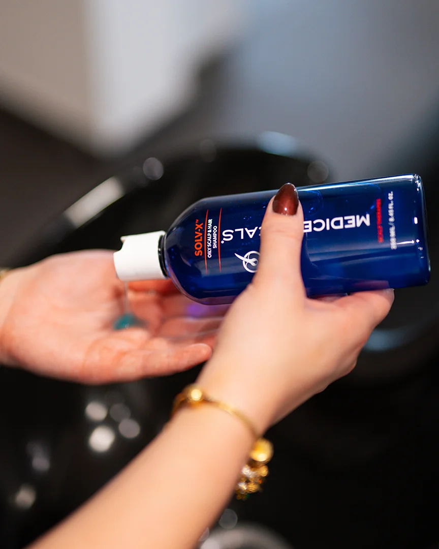 Handen knijpen shampoo uit een blauwe fles boven een zwarte wasbak.