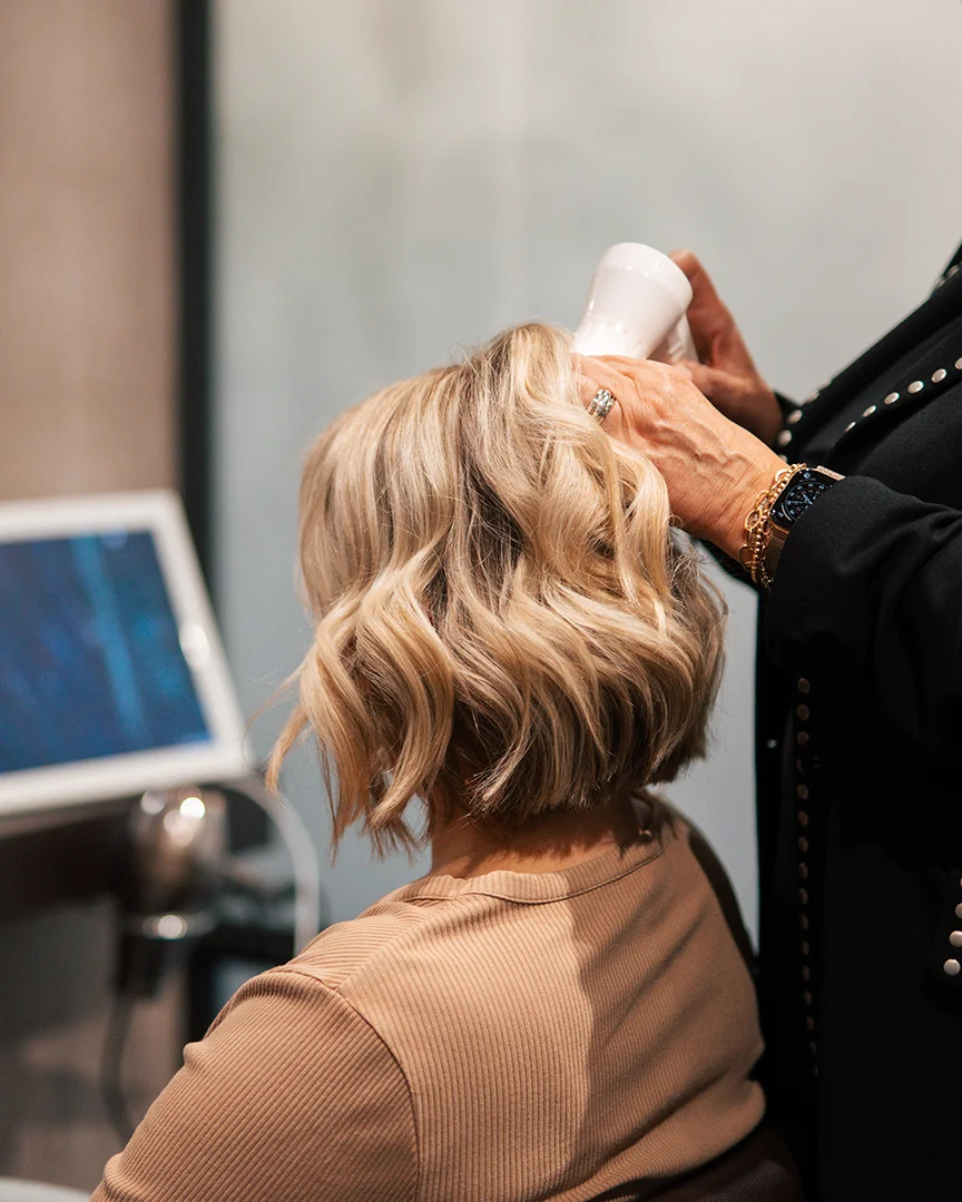Kapper föhnt golvend, blond haar bij een klant met een beige trui.