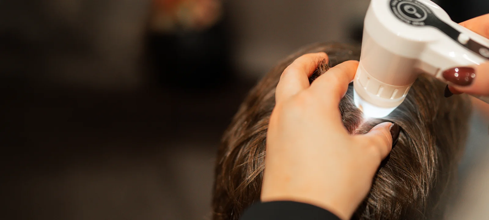 Hand inspecting scalp of dark-haired person using a handheld device with light.