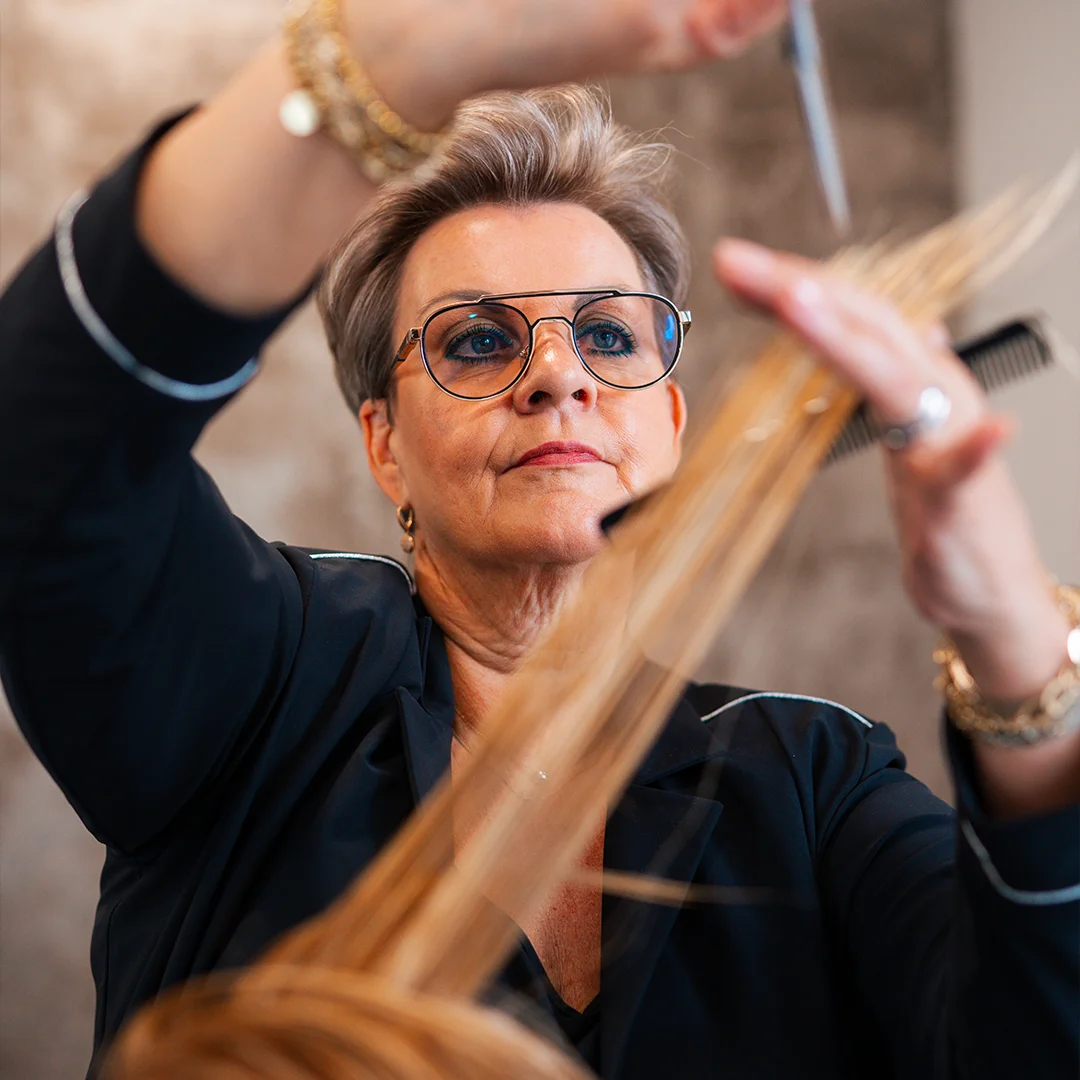 Vrouwelijke kapper met bril knipt geconcentreerd blond haar met schaar en kam.
