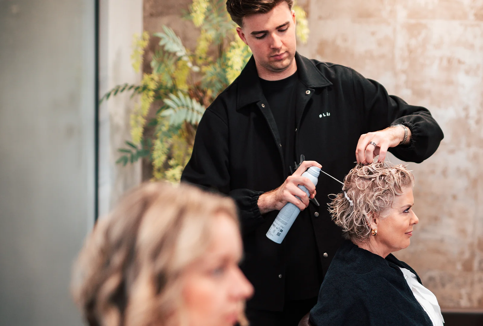 Kapper brengt haarproduct aan op kort, golvend haar van een zittende vrouw in een salon.