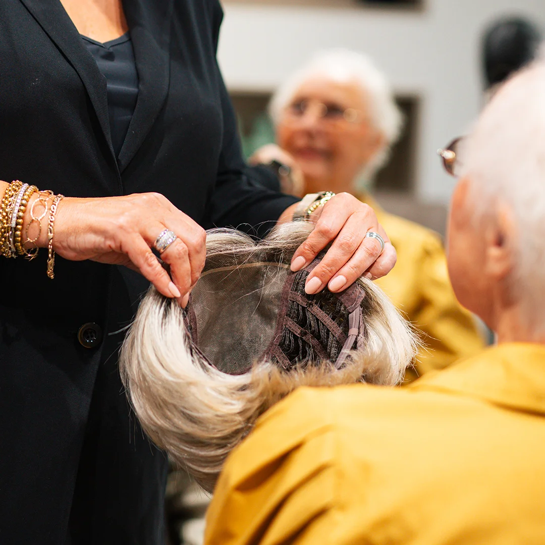 Kapper toont een haarwerk aan een oudere vrouw in geel jack in salonomgeving.
