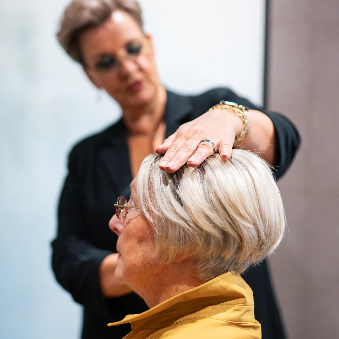 Kapper onderzoekt het hoofd van een oudere vrouw met blond haar en een gele jas.