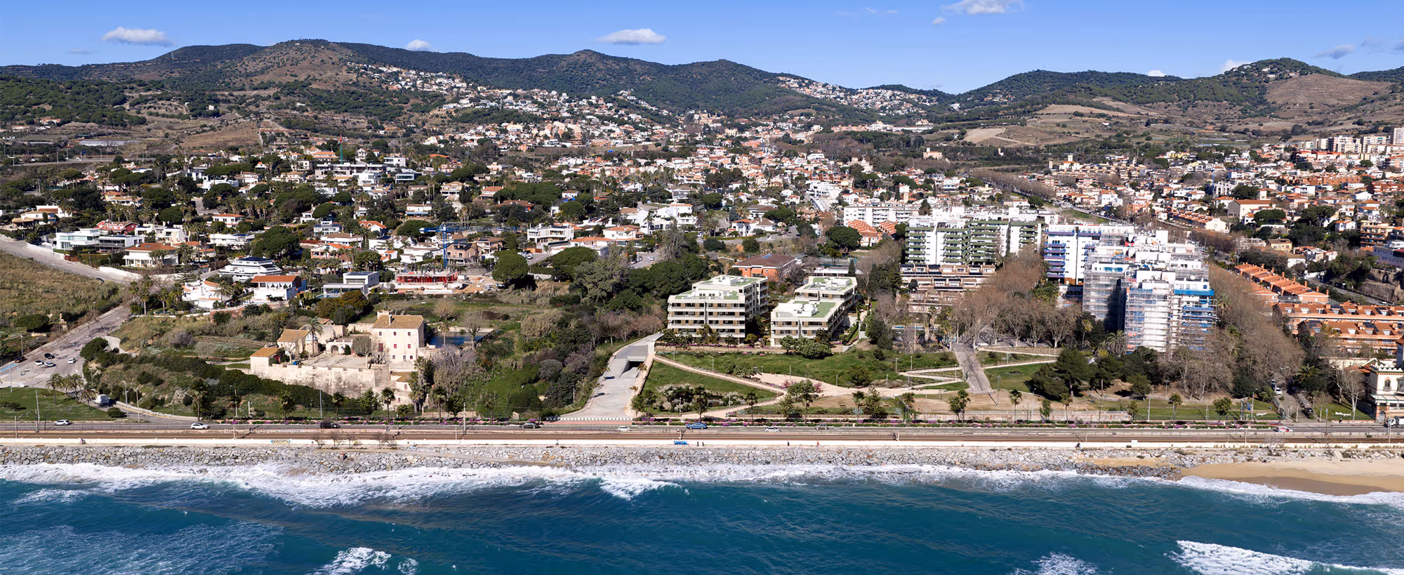 Coastal town with residential buildings, green hills, a beach, and a stone seawall under a clear sky.