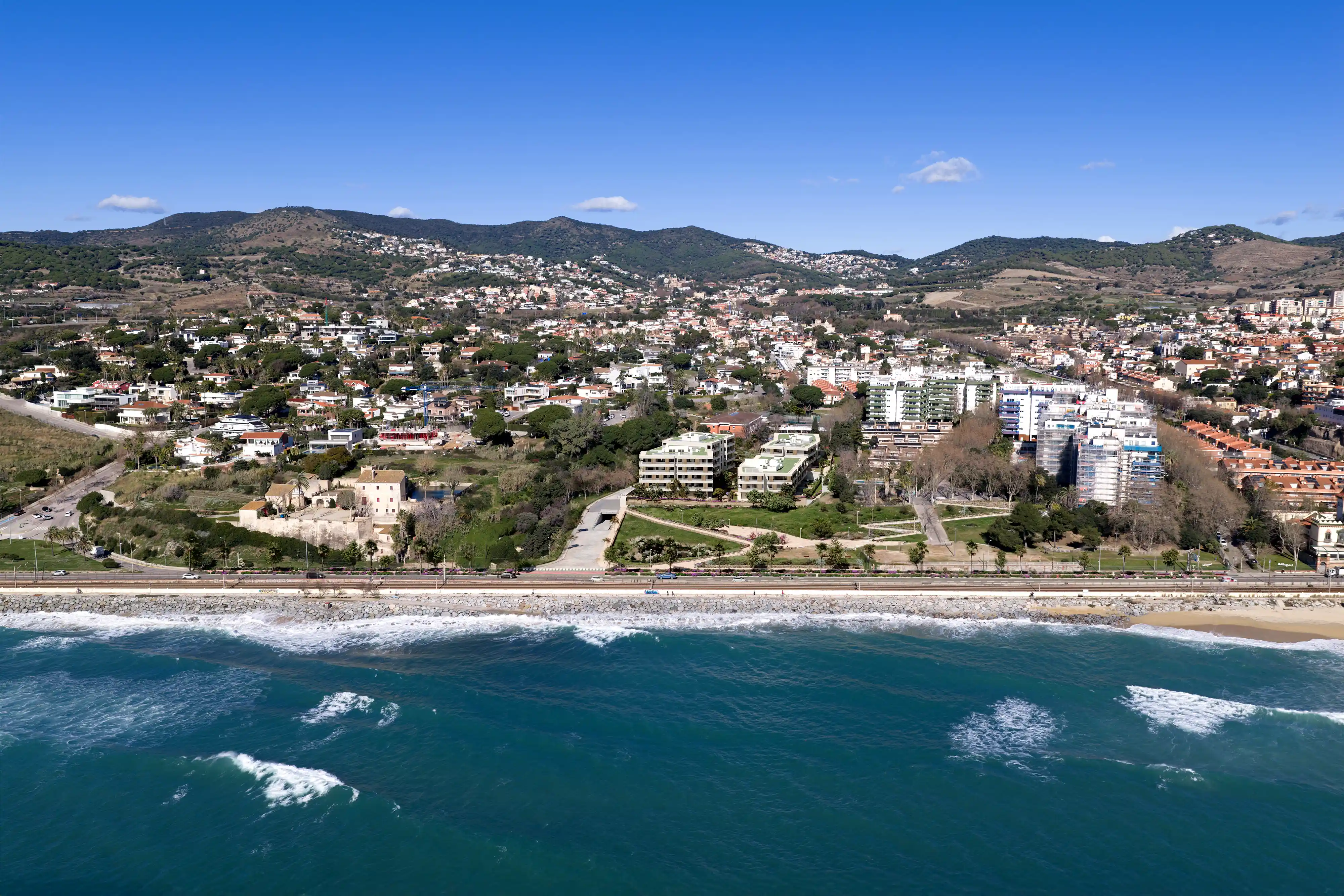 Coastal town with residential buildings, green hills, a beach, and a stone seawall under a clear sky.