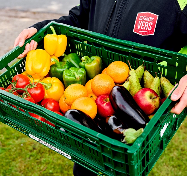 Persoon draagt een groene krat met diverse verse groenten en fruit: gele paprika's, groene paprika's, tomaten, sinaasappels, peren, aubergines en rode appels.