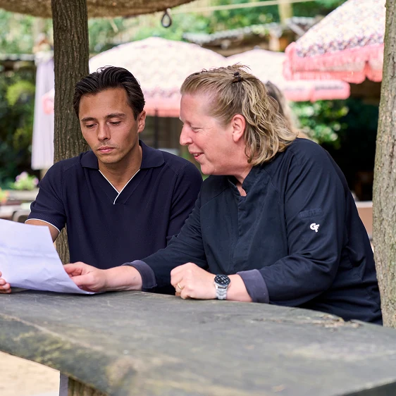Twee mensen zitten buiten aan een houten tafel, de vrouw wijst naar een papieren document dat de man vasthoudt terwijl ze het bespreken.