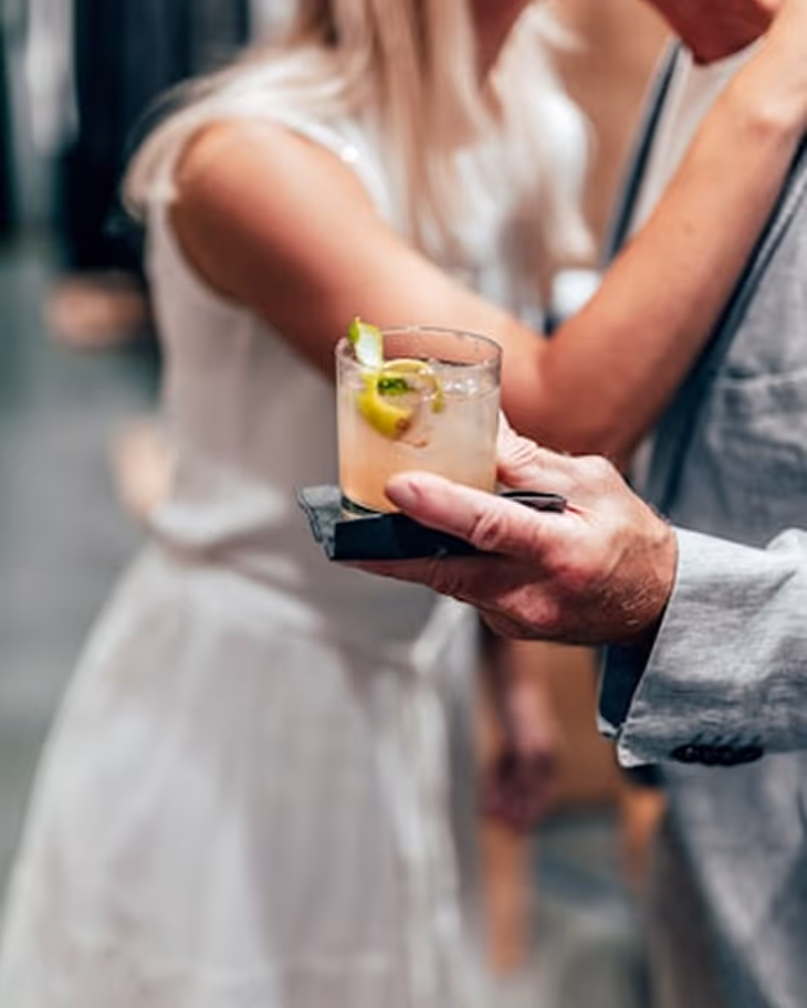 Un homme tenant un verre de cocktail avec un zeste de citron devant une femme en robe blanche floue.