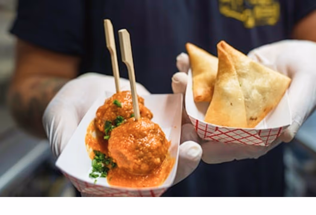 Deux boulettes en sauce avec herbes vertes et deux samoussas dans des plateaux tenus par une personne gantée.