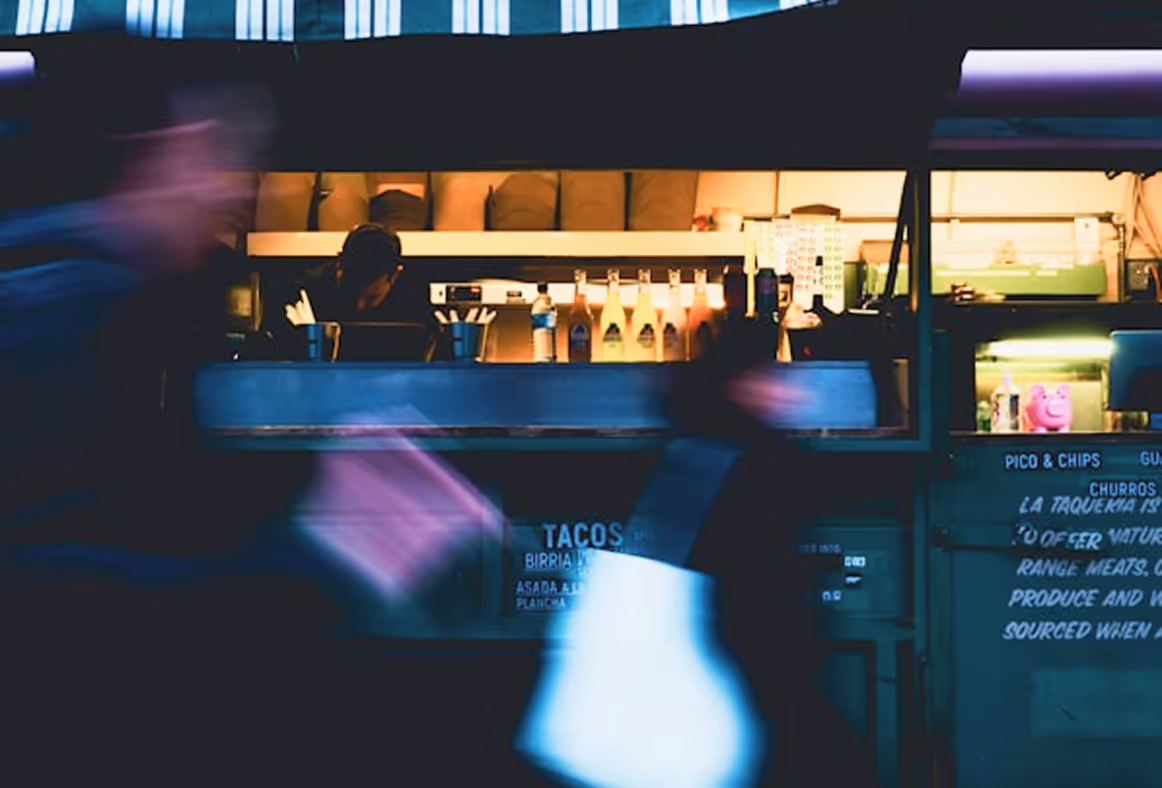 Food truck at night with blurred people walking in front and a worker inside preparing orders.