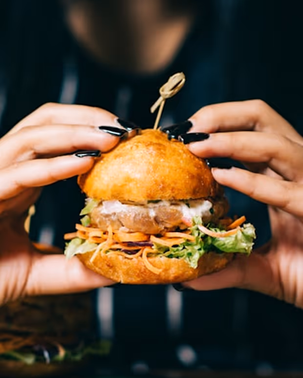 Person tenant un hamburger avec de la laitue, des carottes et une brochette en bois au-dessus.