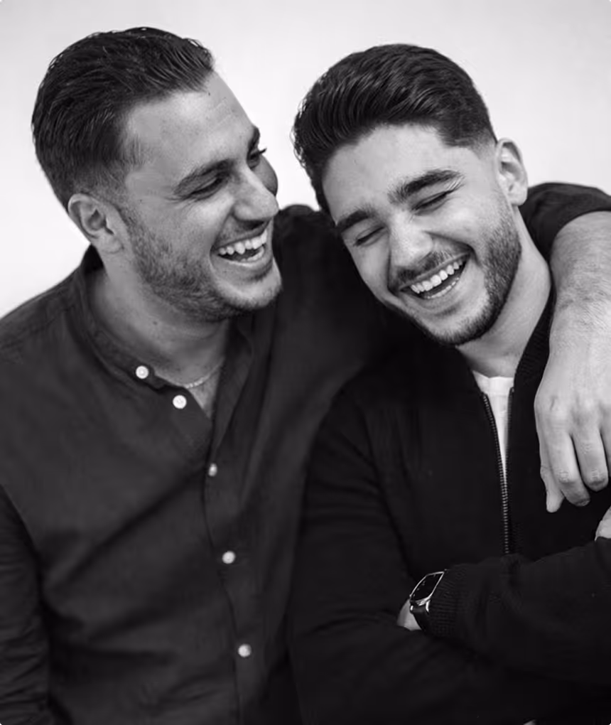 Deux hommes souriants, l'un avec son bras autour de l'autre, partageant un moment joyeux.