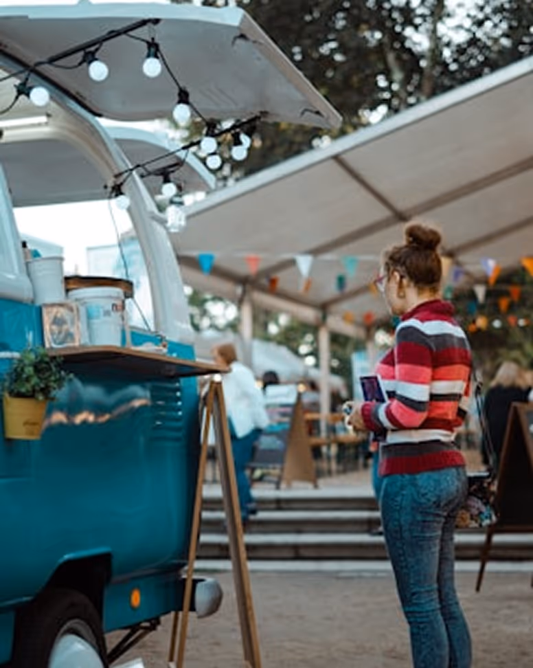 Une femme en pull rayé regarde un camion-restaurant bleu avec guirlande lumineuse lors d'un marché en plein air.