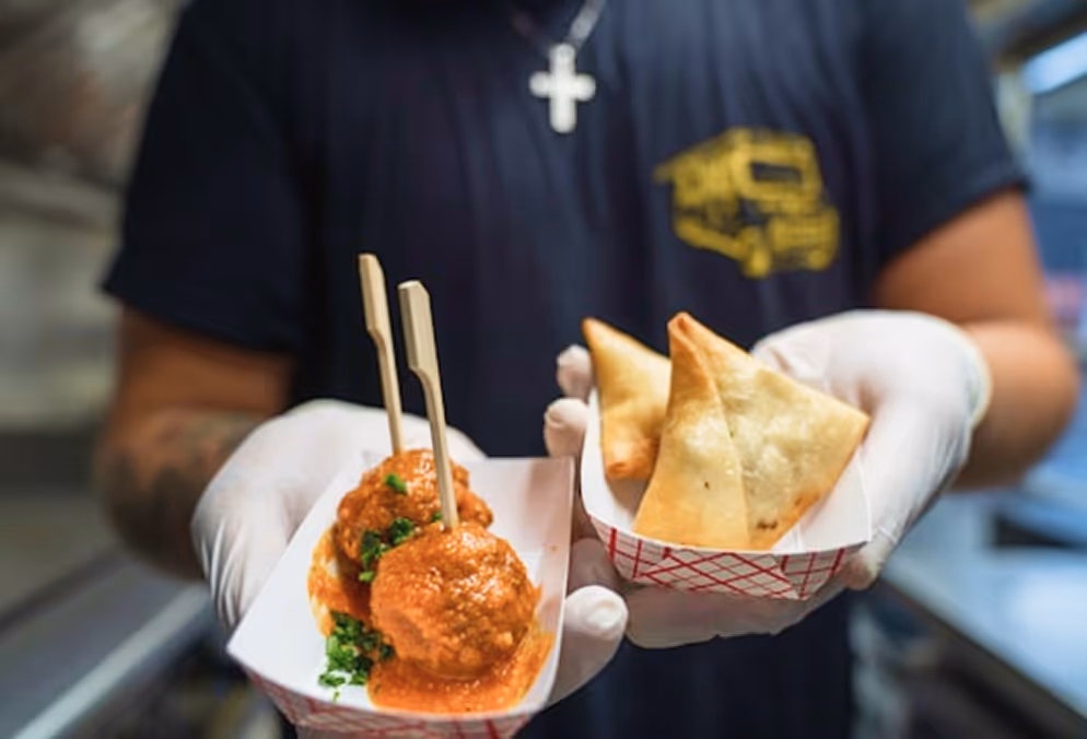 Personne tenant deux plateaux jetables avec des boulettes en sauce et des samoussas frits.