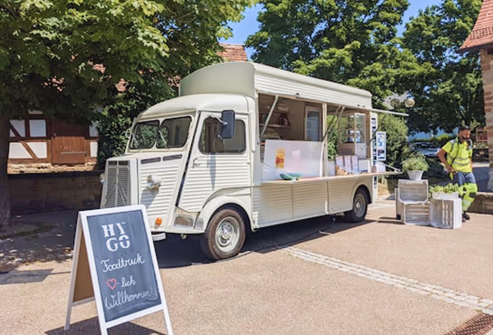 Food truck blanc stationné sous un arbre avec panneau d’accueil et homme en jaune à côté.