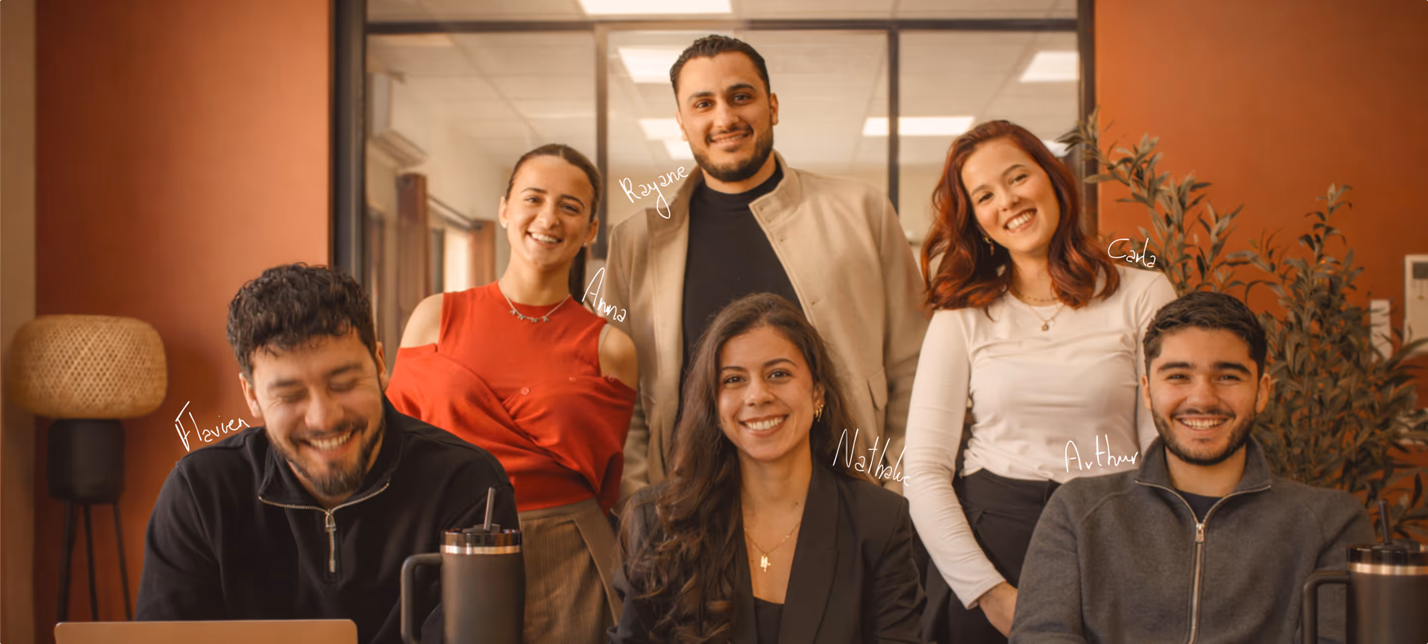 Groupe de six jeunes souriants en tenue décontractée dans un bureau moderne avec mur orange.