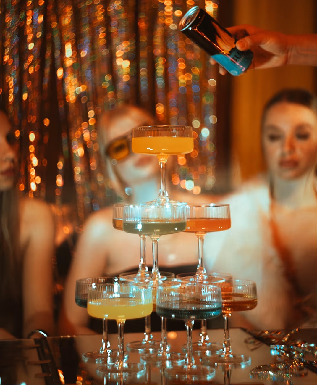 Verres de cocktail colorés empilés en pyramide avec une main tenant une canette au-dessus.