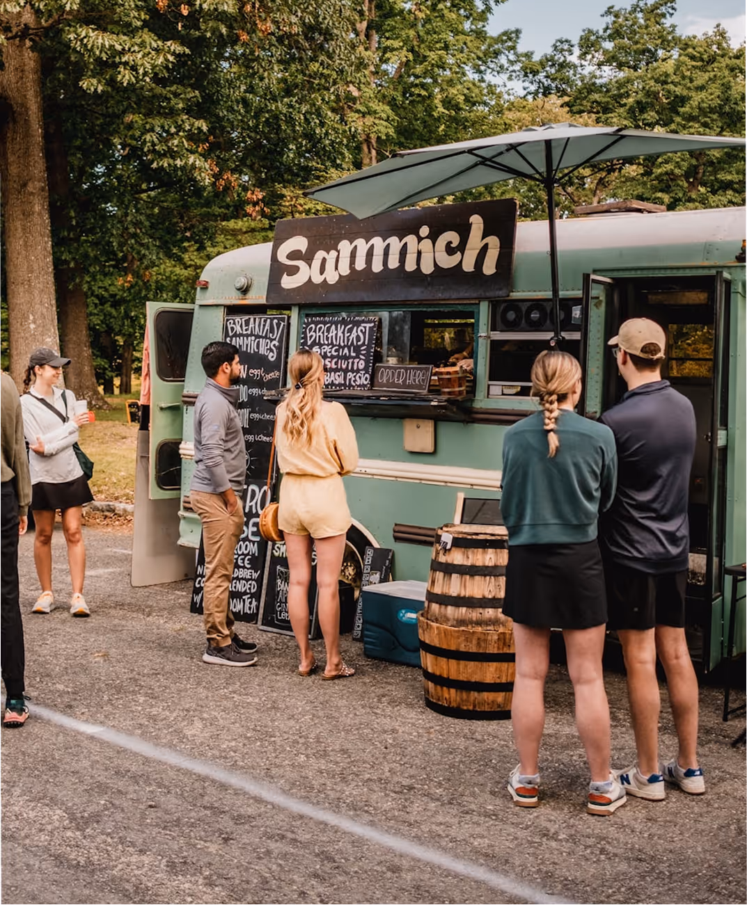 Des personnes font la queue devant un food truck vert nommé Sammich dans un parc.