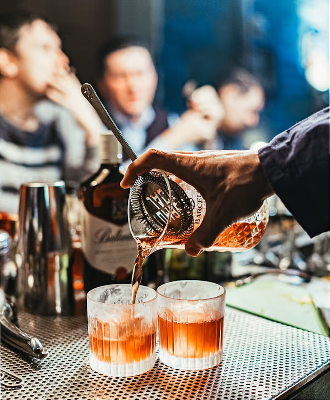 Une main verse un cocktail brun dans un verre à whisky au bar avec des personnes en arrière-plan floues.