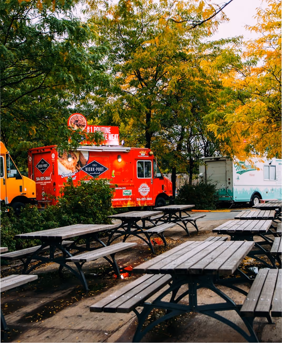 Tables de pique-nique vides en bois avec camions de nourriture rouge et jaune en arrière-plan sous des arbres d'automne.