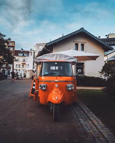 Petit tuk-tuk orange garé sur une route urbain devant une maison sous un ciel bleu clair.