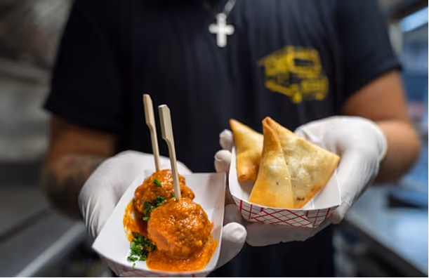 Deux boulettes en sauce orange et triangles frits servis sur plateaux blancs par une personne gantée.