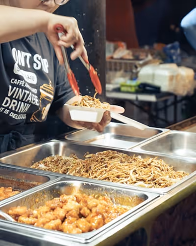 Personne servant des nouilles dans un plateau parmi plusieurs plats chauds en buffet.