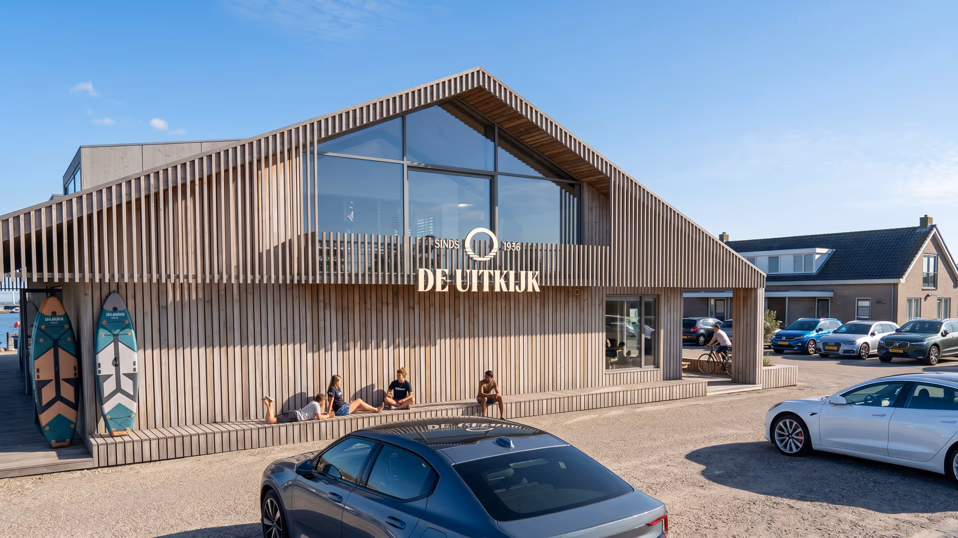 Modern houten gebouw De Uitkijk met mensen die buiten op het bankje zitten en geparkeerde auto's op een zonnige dag.