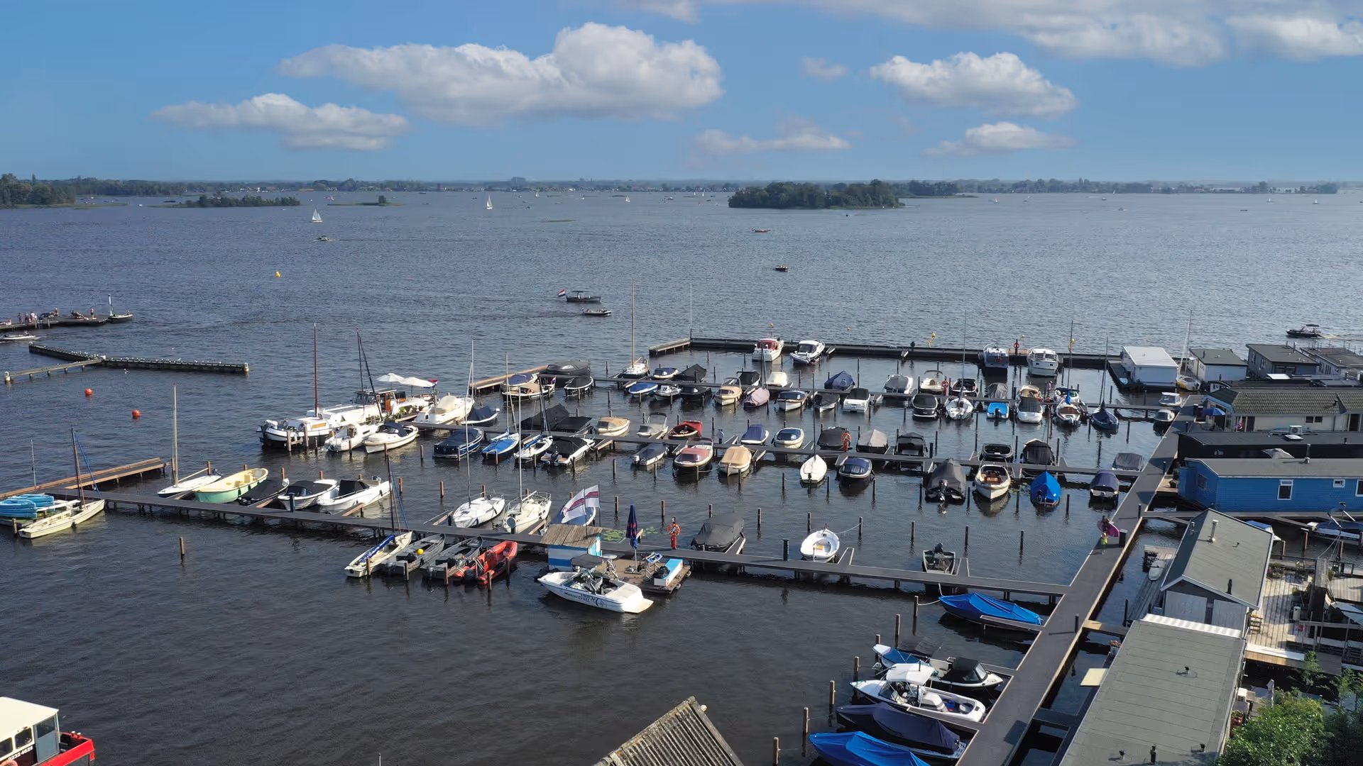 Luchtfoto van een jachthaven met verschillende kleine boten en zeilboten aangemeerd aan steigers onder een blauwe hemel met enkele wolken.