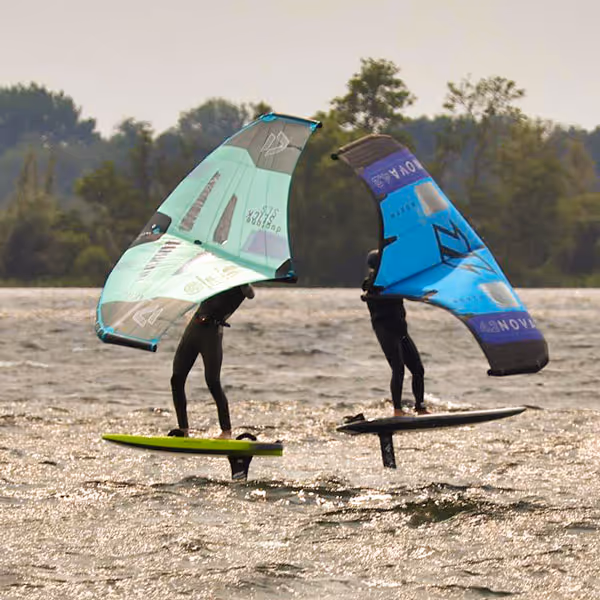 Twee mensen wingsurfen op foilboards met groene en blauwe vleugels boven het water.