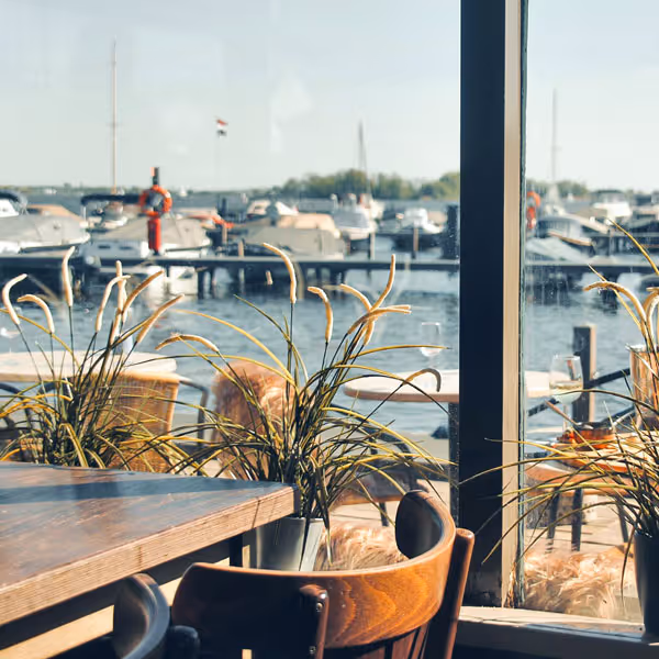Gezellige cafésetting met houten tafels, stoelen en planten, uitzicht op een havendok met aangemeerde boten.