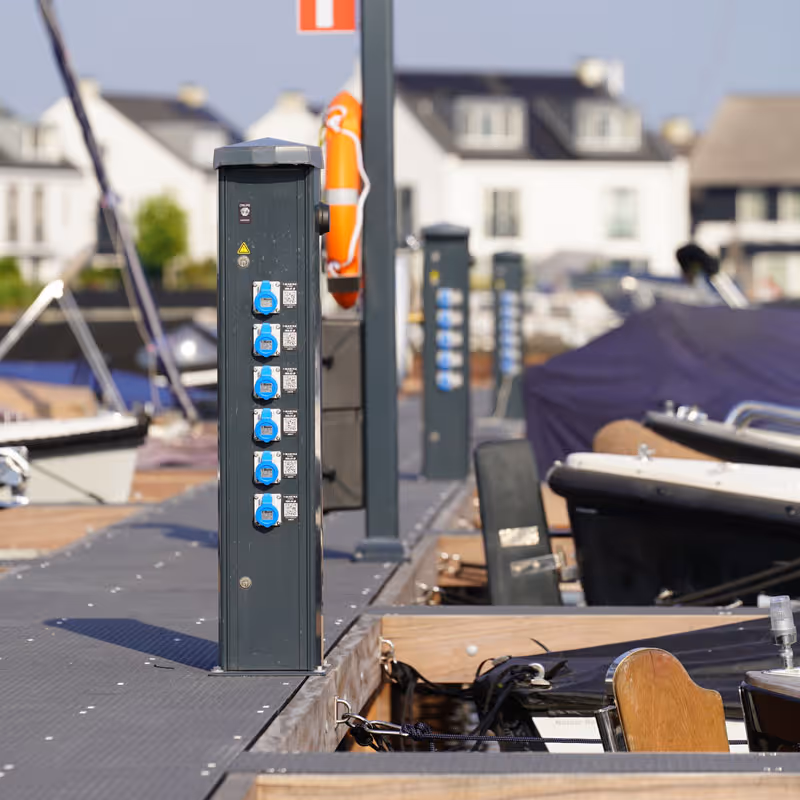 Steiger met stroompalen en aanlegplaatsen voor boten in een jachthaven met huizen op de achtergrond.