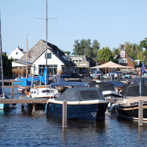 Woonboten en zeilboten liggen aangemeerd bij een rustige haven met huisjes en bomen op de achtergrond onder een blauwe lucht.