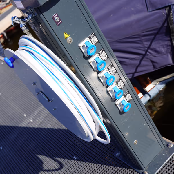 Elektrische stroomvoorziening met vijf blauwe stopcontacten en een opgerolde witte waterslang aan een donkere paal op een steiger.