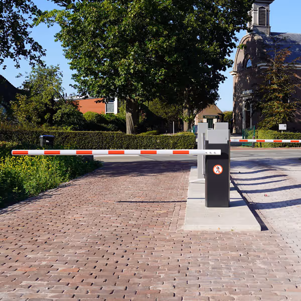 Automatische slagbomen bij een toegang tot een weg met bakstenen bestrating en bomen in de achtergrond.