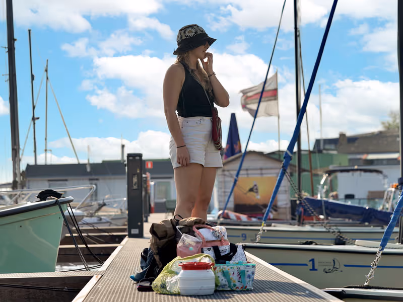 Vrouw in zwarte top, witte korte broek en hoed staande op een steiger met tassen en spullen rondom haar, naast aangemeerde boten onder een deels bewolkte lucht.