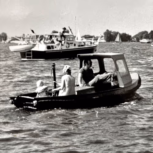 Zwart-wit foto van een kleine motorboot met drie mensen op een druk vaarwater met zeilboten op de achtergrond.
