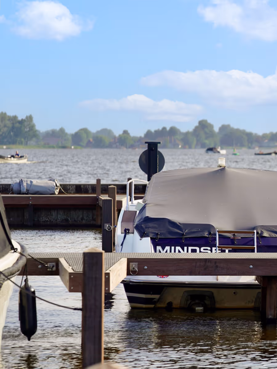 Motorboot met grijs zeil met opschrift Mindset afgemeerd aan een houten steiger op een meer met blauwe lucht en bomen op de achtergrond.
