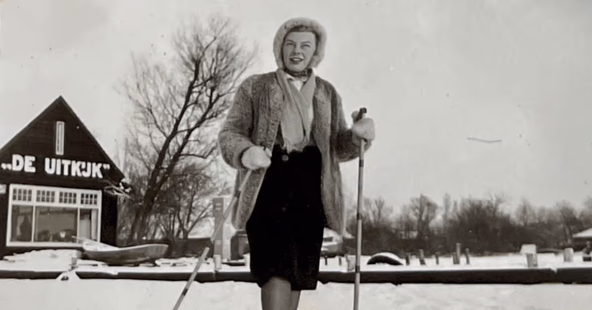 Zwart-witfoto van een vrouw in winterkleding met ski's en stokken staand op sneeuw met een gebouw 'De Uitkijk' op de achtergrond.