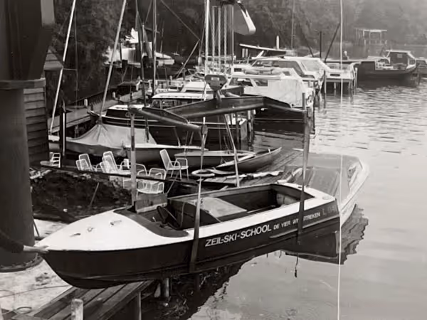 Zwart-witfoto van een kleine boot met de tekst 'Zeil-Ski-School De Vier Winden' aangemeerd bij een dock met meerdere boten en stoelen aan het water.