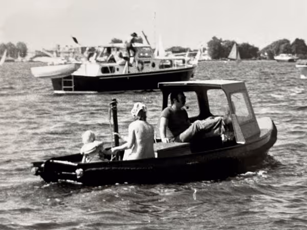 Zwart-wit foto van een klein bootje met drie personen, waaronder een kind en een vrouw met een hoofddoek, varend op het water met grotere boten en zeilboten op de achtergrond.