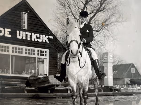 Zwarte-witfoto van een man in ruiterkleding op een wit paard voor een gebouw met het bord 'DE UITKIJK'.