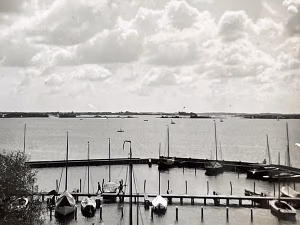 Zwart-wit foto van een haven met zeilboten en stille wateren onder een bewolkte hemel.