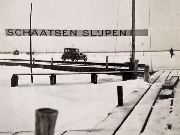 Oude zwart-witfoto van een besneeuwde houten steiger met een vintage auto en een persoon in de verte, met een bord waarop staat 'Schaatsen Slijpen'.