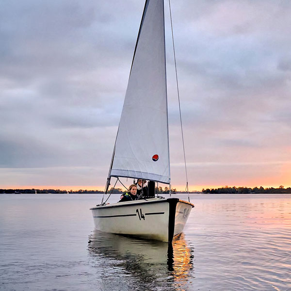 Zeilboot met twee personen vaart over kalm water bij zonsondergang.