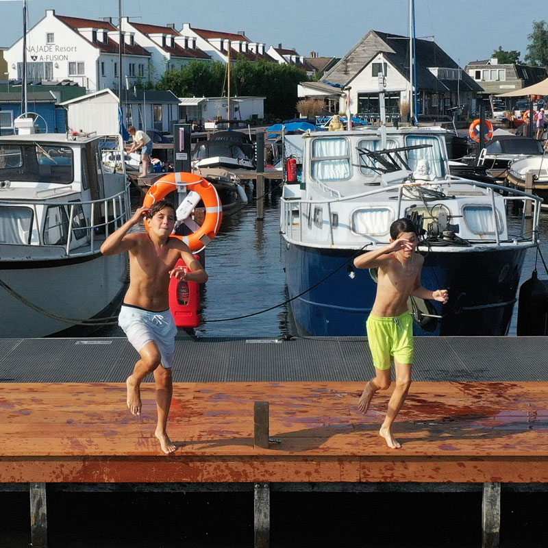 Twee jongens in zwemkleding rennen over een natte houten steiger bij een haven met boten.