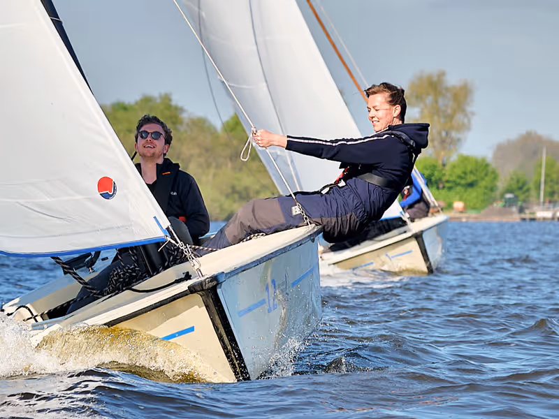 Twee mensen zeilen op kleine boten op een kalme waterplas onder een blauwe lucht.