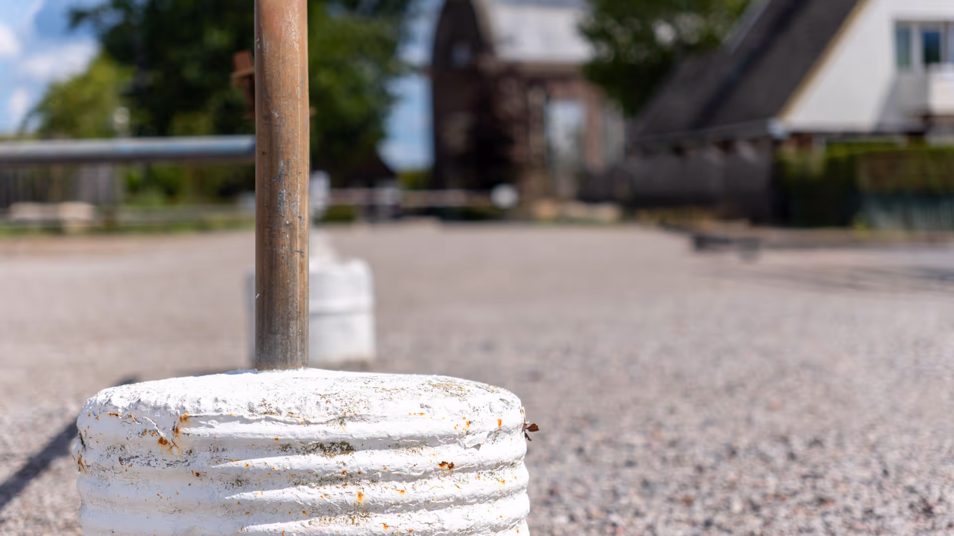 Witte metalen parkeerpaal met roest en een grindpad met onscherpe gebouwen op de achtergrond.