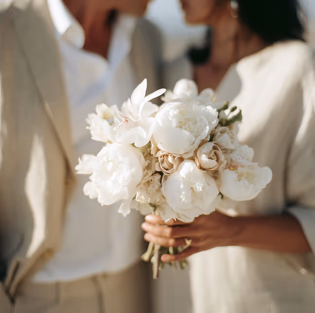 Close-up of a couple holding a bouquet of white and pale peach flowers, both dressed in light beige clothing.