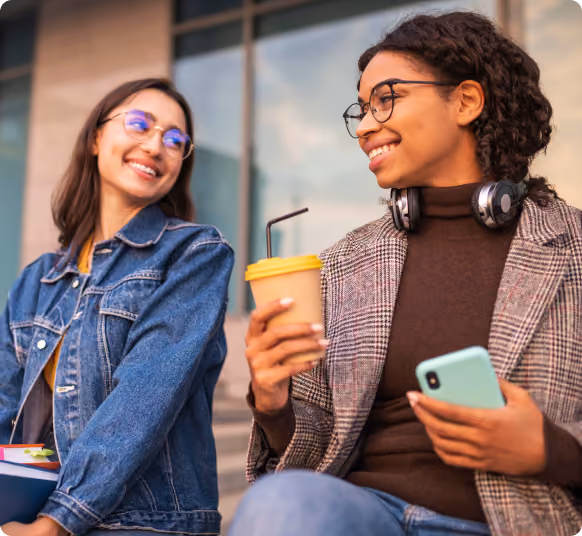 Duas alunas sorrindo na entrada da faculdade, uma segurando o celular e um copo de café, e a outra com livros e uma mochila.